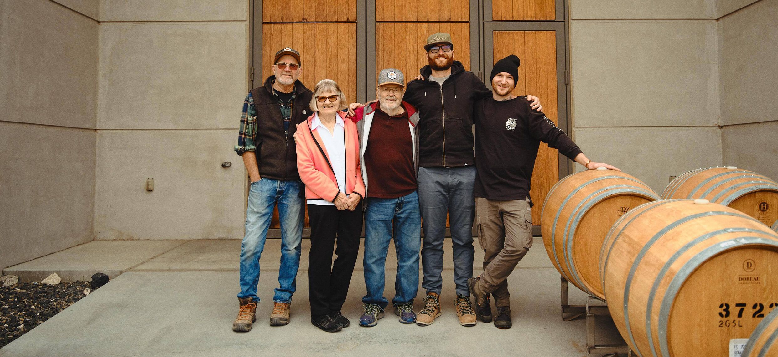 The Kiona family and team standing arm in arm in front of the winery's wooden cellar doors, beside a row of oak barrels
