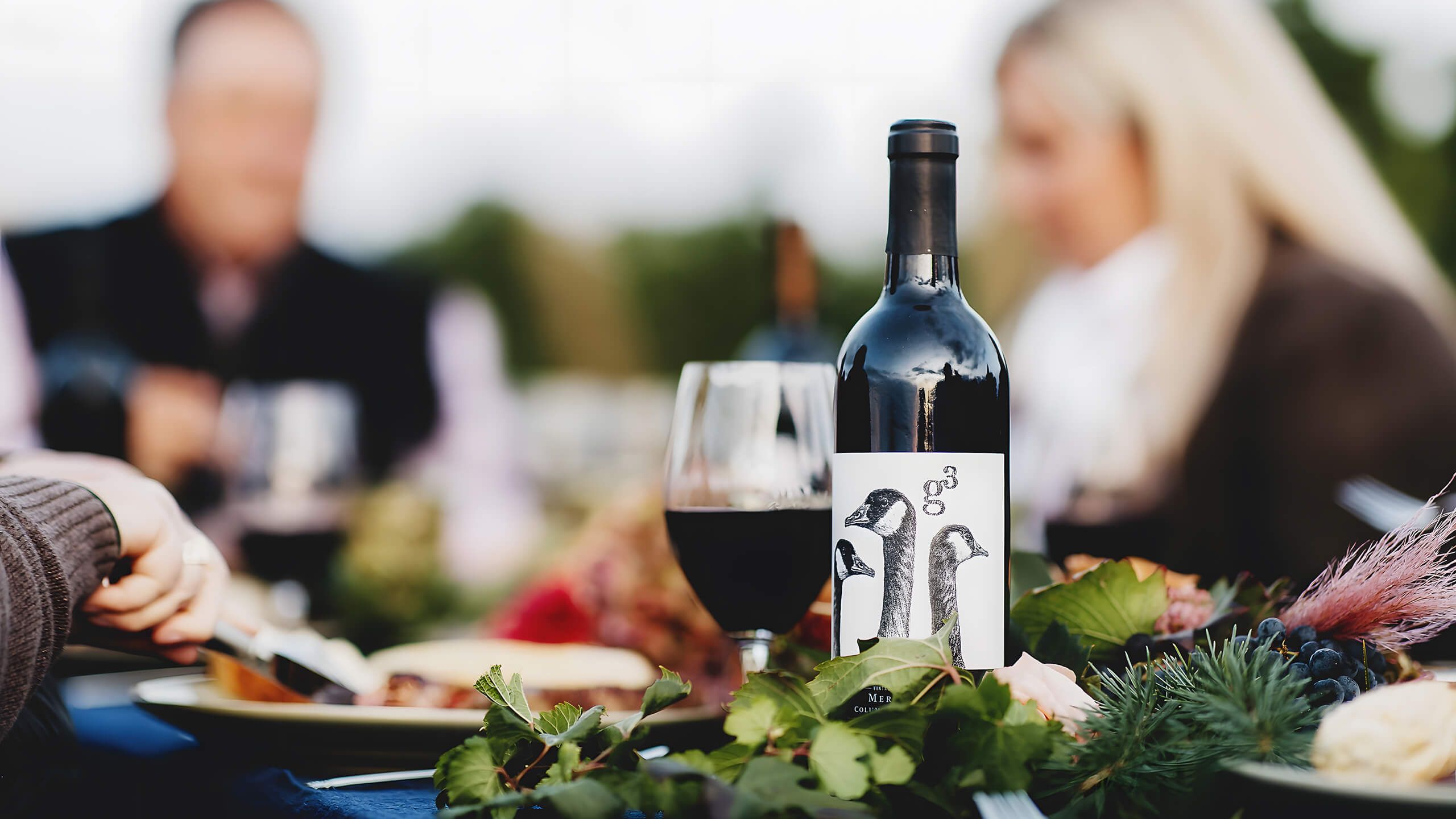 A bottle of Goose Ridge g3 Merlot, its label drawn with Canada geese, beside a glass of red wine on a dinner table set among greenery