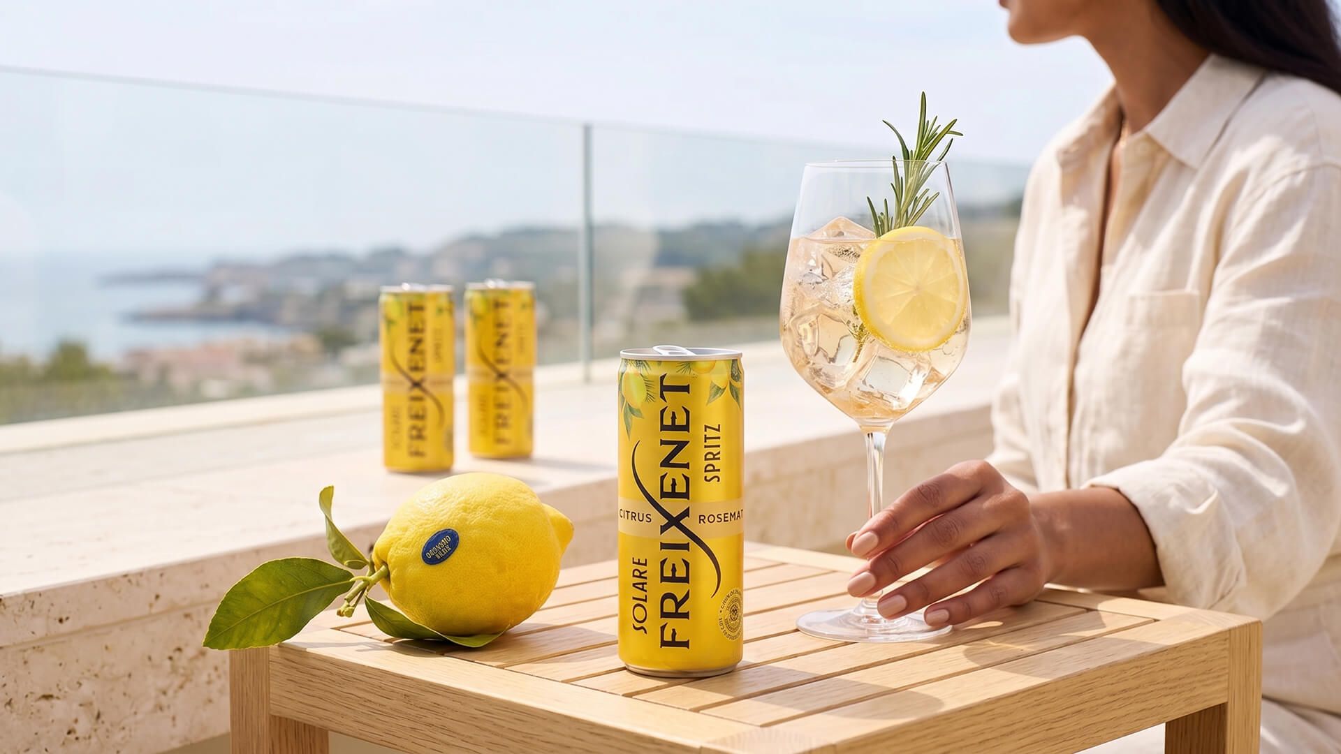 Freixenet Solare Spritz can on a teak side table between a fresh lemon and a poured spritz, a guest's hand on the glass, sunlit sea terrace behind